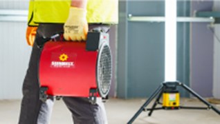 Man on a working site carrying a small red portable heater 
