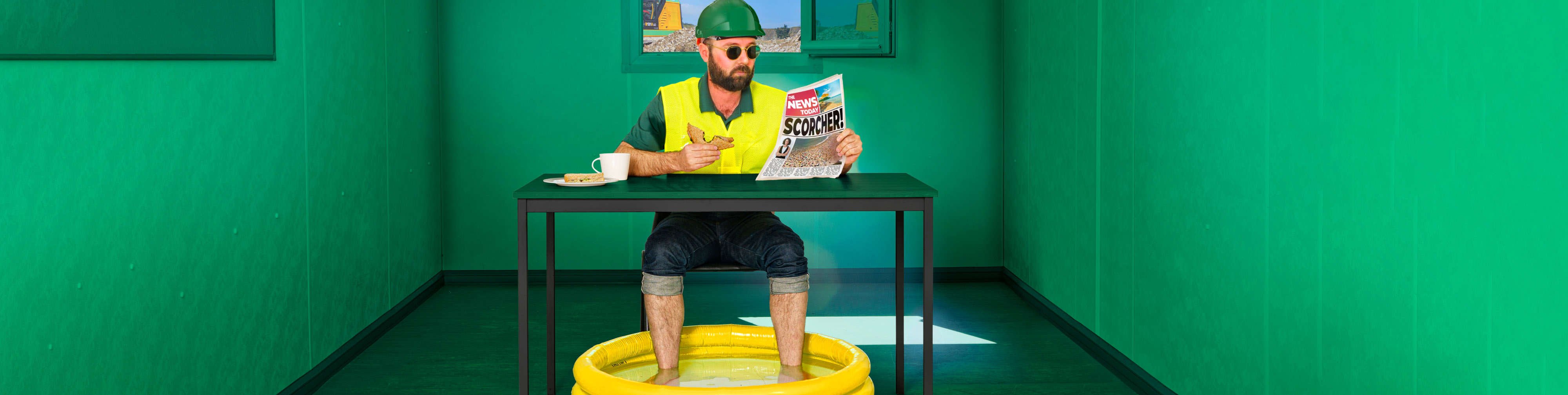 Construction worker sat eating lunch at a table trying to keep cool with there feet in a paddling pool
