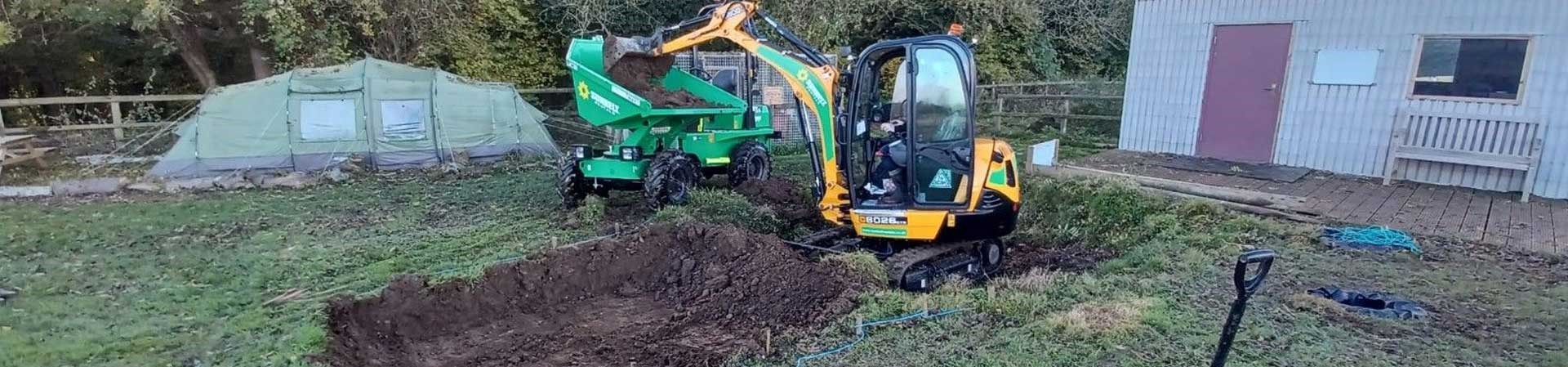 Mini Digger Unloading Dirt Into A Dumper On Patch Of Grass Worksite