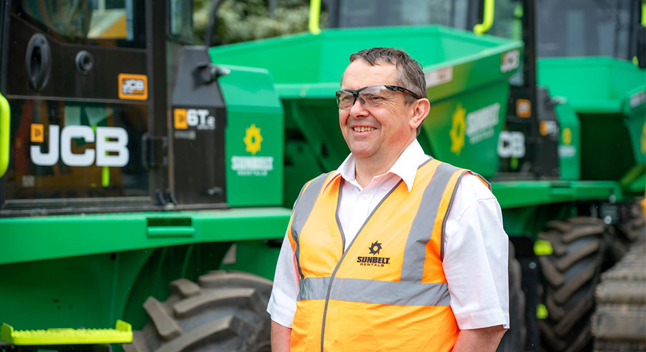 Stuart Sunbelt Rentals Worker Stood In Woker Yard Next To Two Sunbelt Dumpers Wearing A Hi Vis And Safety Glasses