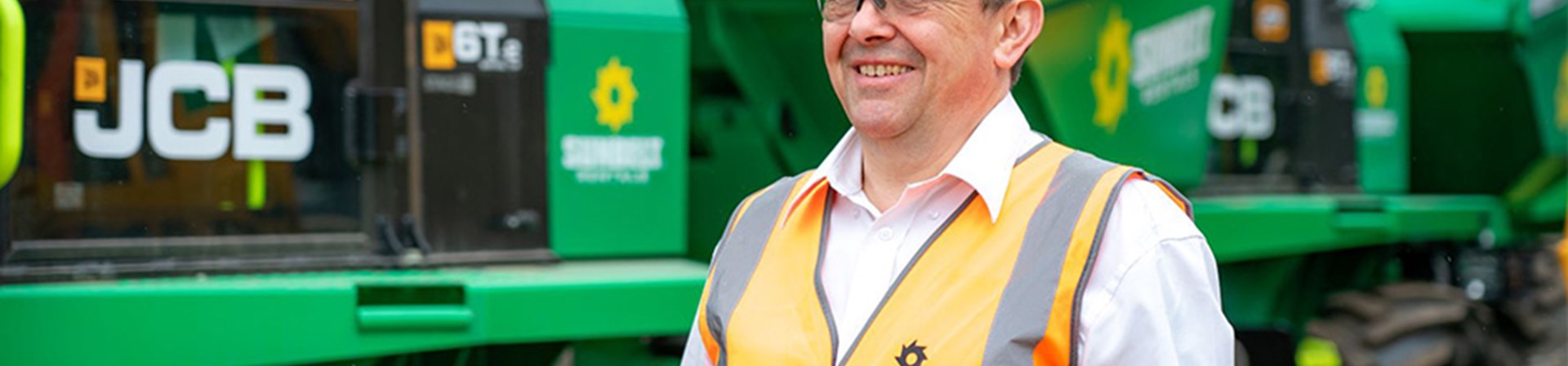 Stuart Sunbelt Rentals Worker Stood In Woker Yard Next To Two Sunbelt Dumpers Wearing A Hi Vis And Safety Glasses